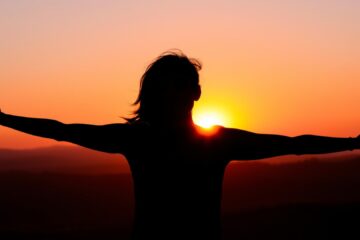 A_dark_silhouette_of_a_woman_with_her_arms_stretched_wide_standing_against_a_vibrant_sunset.