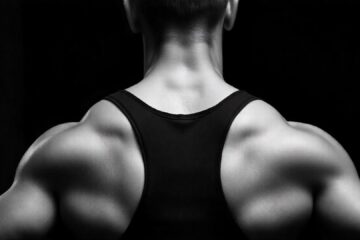 The image shows the back of a fit man, highlighting her muscular definition. He is wearing a black sports bra that exposes her upper back and shoulders. The focus is on her strong, toned muscles, especially her shoulder blades, upper back, and arms, which are well-defined. The background is dark and blurred, placing full emphasis on her physique. The lighting is soft but directed in a way that accentuates the contours of her muscles, making the image feel dramatic and powerful.